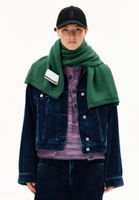 Personne portant une casquette noire, une veste en velours côtelé bleu foncé et un pantalon, un pull en maille violet et une écharpe verte avec une étiquette blanche "ARMEDANGELS".