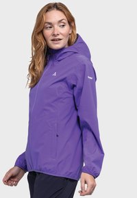 Frau in einer lila Kapuzenjacke, die nach links schaut, mit entspannten Händen an den Seiten vor einem schlichten, hellen Hintergrund.