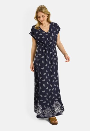 Mujer vestida con un maxi vestido azul marino con estampado de cachemira blanco y mangas cortas, combinada con sandalias de plataforma marrones, de pie sobre un fondo blanco.