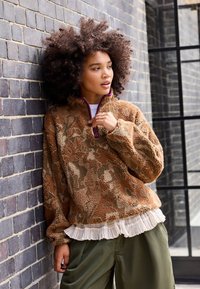 Bruin met patroon fleece pullover met een ritskraag, voorzien van paarse accenten. Gelaagd met een frêle witte shirt en gecombineerd met groene broeken.