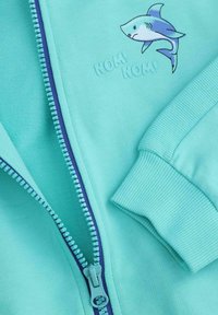 Jasnoniebieska bluza z wiązaniem na suwak i ściągaczami przy rękawach. Posiada grafikę rekina oraz wytłoczony tekst "NOM! NOM!" w pobliżu suwaka. Miękka w dotyku.