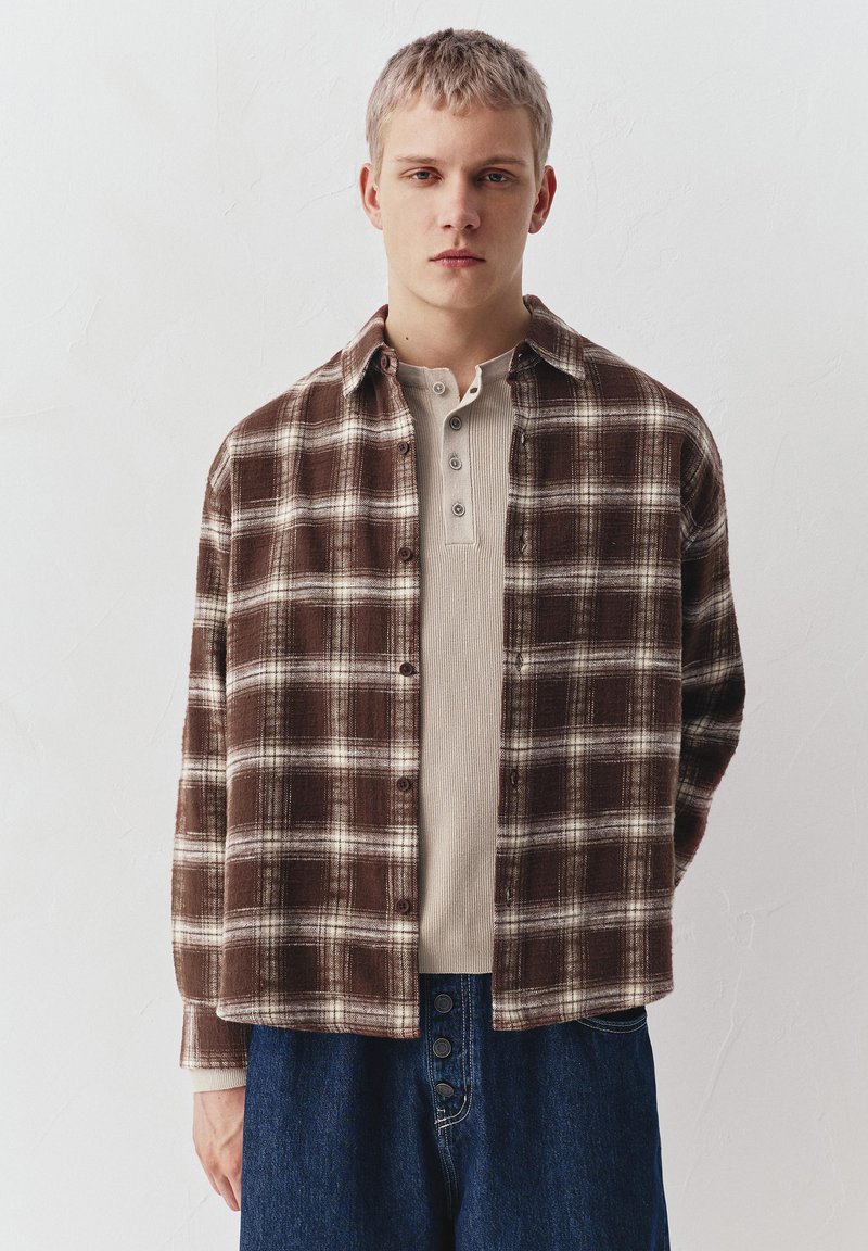 Junger Mann mit kurzen blondem Haar, der ein braun-weiß kariertes Hemd über einem beigen Henley trägt, kombiniert mit dunkelblauen Jeans.