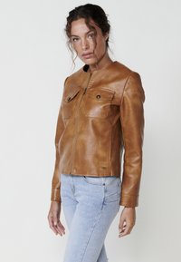 Giacca in pelle marrone con chiusura a zip, colletto alto e due tasche sul petto. Presenta una texture liscia, un design aderente e dettagli del marchio discreti.