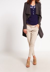 Giacca aperta grigio scuro sopra una blusa blu navy con scollatura floreale, pantaloni a sigaretta color crema e sandali beige perforati.