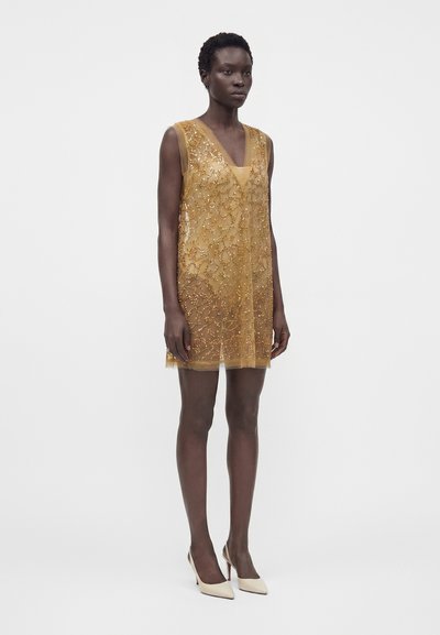 Femme portant une robe dorée sans manches en tissu transparent avec des ornements floraux en perles, assortie à des talons blancs à bout pointu, debout devant un fond uni.