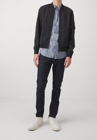 Giubbotto bomber nero, camicia a righe azzurra, jeans scuri e sneakers bianche. Tessuto liscio e vestibilità casual con chiusura a zip e polsini a costine.