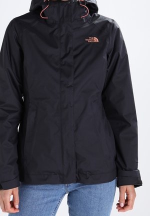 Personne portant une veste imperméable noire North Face avec capuche et cordons roses, associée à un jean bleu.