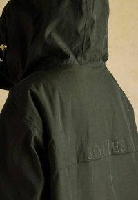 Mørkegrøn vandtæt jakke med hætte. Har et struktureret stof og et subtilt "JOULES"-logo syet på ryggen.