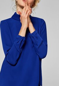 Blusa blu a maniche lunghe con colletto falso, realizzata in un tessuto leggero e liscio, con polsini abbottonati e una vestibilità rilassata.