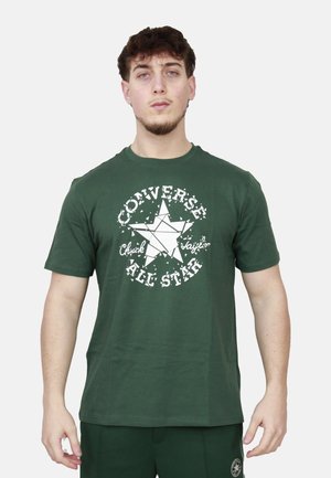 Giovane uomo con capelli corti e ricci che indossa una t-shirt verde Converse All Star e pantaloni verdi abbinati, in piedi davanti a uno sfondo neutro.