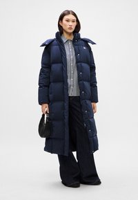 Námornícky modrý, dlhý puff coat s prešívaným dizajnom, zapínaním na gombíky a širokým golierom. Nosený cez pruhované tričko a v kombinácii s tmavými džínsami.