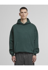 Mørkegrønn hettegenser med snor i hetten, oversized passform, frontlomme og ribbede mansjetter, laget av myk stoff med en glatt tekstur.