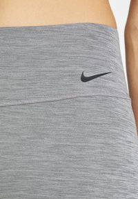 Grå højtaljede leggings lavet af tekstureret stof, med et sort Nike-logo og synlig søm langs taljebåndet.