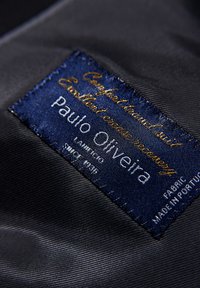 Niebieska etykieta z tkaniny z złotym haftowanym tekstem; zawiera napisy "Paulo Oliveira", "Excellence" oraz "Made in Portugal", prezentując gładką teksturę i haft.