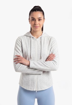 Jonge vrouw met donker haar in een paardenstaart draagt een lichtgrijze hoodie met rits en blauwe leggings, staat zelfverzekerd met de armen over elkaar.