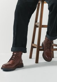 Braune Leder-Schnürstiefel mit rundlicher Spitze, Gummisohle und Knöchelhöhe, kombiniert mit hochgekrempelten schwarzen Jeans, die auf einem Holzstuhl ruhen.