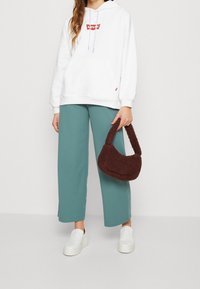 Bílá mikina s červeným logem, teal široké kalhoty, bílé nazouvací boty a hnědá chlupatá kabelka. Neformální outfit s jemnými texturami.