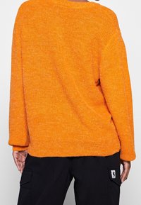 Oranger gestrickter Pullover mit lockerer Passform, weitem Halsausschnitt und langen Ärmeln. Strukturierter Stoff und Saum, der auf den Hüften endet.