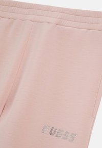 Helles rosa Jogginghosen aus weichem Stoff, mit einem strukturierten Bund und einem silbernen Logo, das auf dem linken Bein gedruckt ist.