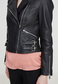 Svart läderbikerjacka med silverfärgad dragkedjor och dubbla sidspännen, bärs över en rosa blus och svarta byxor.