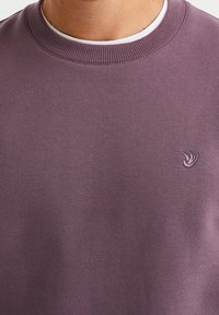 Lila sweatshirt i bomullsmix, med ribbstickad rund halsringning och liten fågallogotyp broderad på vänster bröst. Slät textur.