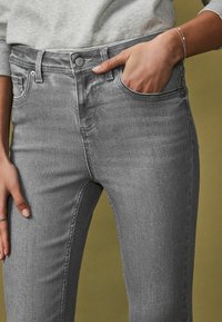 Mujer con jeans de mezclilla gris y un suéter gris claro, con la mano derecha en el bolsillo delantero y una pulsera de plata en la muñeca.