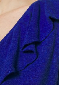 Blått stickat tyg med en mjuk textur, som har en volangdesign längs halslinjen och fångar olika nyanser av blått genom stickningen.