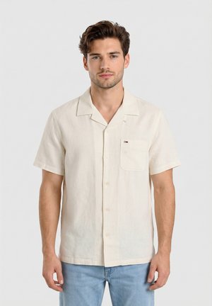 Uomo con camicia crema a maniche corte abbottonata con taschino sul petto e jeans azzurri chiaro, in piedi davanti a uno sfondo chiaro e uniforme.