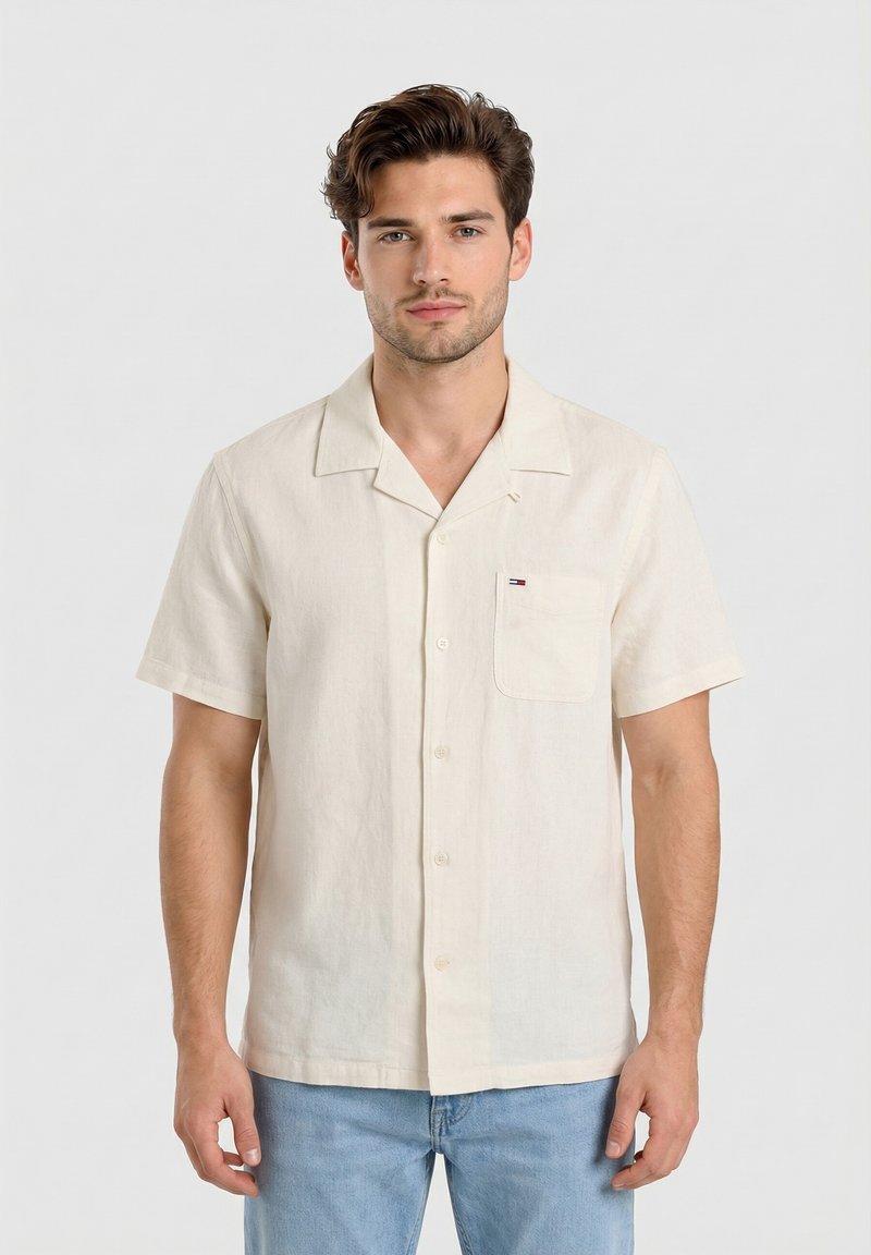 Homme portant une chemise crème à manches courtes boutonnée avec poche poitrine et un jean bleu clair, debout devant un fond clair uni.