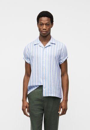 Mężczyzna o krótkich, kręconych włosach, ubrany w jasnoniebieską i białą koszulę w pionowe paski z krótkim rękawem oraz ciemnozielone spodnie, stojący spokojnie.