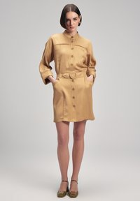 Vestido camisero color caqui con cuello, frente abotonado y bolsillos laterales. Incluye un cinturón a juego y tiene una textura suave, similar a la del satén.