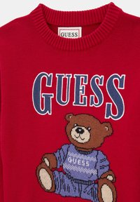 Maglione rosso con un orso di maglia marrone che indossa un outfit blu con la scritta "GUESS." Collo a coste e un design grafico distintivo.