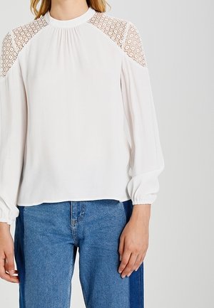 Kobieta ubrana w białą bluzkę z długim rękawem z koronkowymi panelami na ramionach i wysokim dekoltem, zestawioną z dżinsami w dwóch odcieniach niebieskiego.