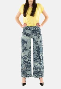 Pantalones de pierna ancha en una tela con tintes azules que presenta un detallado estampado floral y de paisajes. Combinados con una blusa amarilla sencilla y tacones negros.