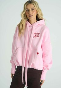 Růžová mikina s kapucí a přední kapsou, zdobená červenou výšivkou "Hot Girls Go To Pilates", vyrobená z jemného materiálu s volnějším střihem.