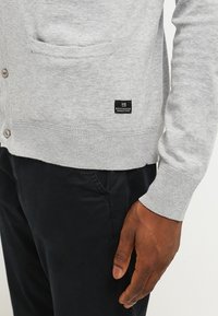 Cardigan de punto gris con un bolsillo, que presenta cierres de botón y puños acanalados. Etiqueta de logo negra en la parte inferior izquierda. Llevado sobre pantalones oscuros.