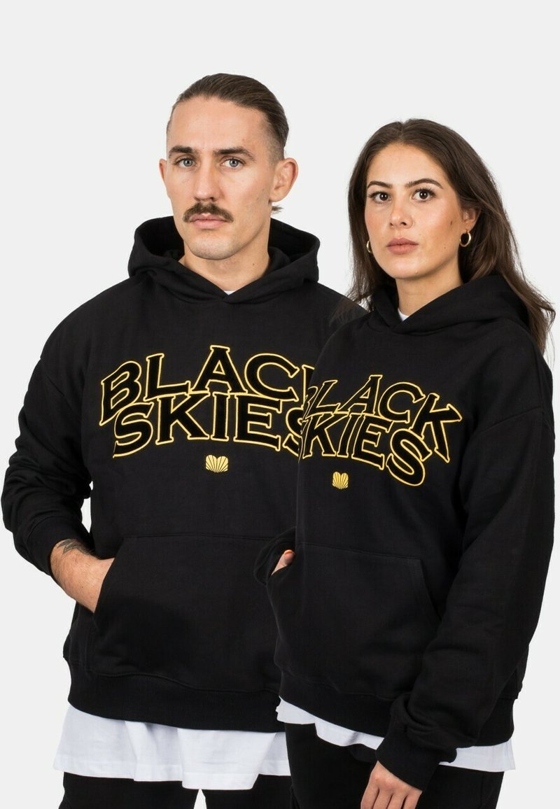 Black hoodie made of cotton featuring large yellow text “BLACK SKIES.” Includes a front pocket and ribbed cuffs and hem. Simple design.