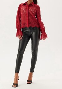 Blouse en dentelle rouge avec manches longues évasées, associée à un legging en similicuir noir et des talons ouverts noirs. La tenue comprend un col et des détails en tulle.