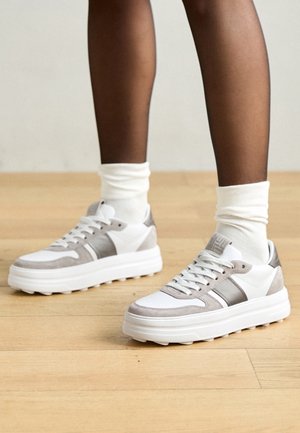 Personne portant des baskets blanches et grises avec des semelles épaisses et des chaussettes blanches, debout sur un sol en bois clair.