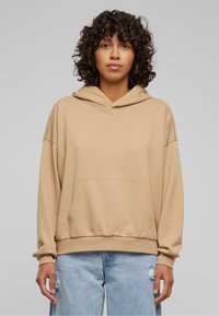Sudadera beige con capucha y bolsillo canguro, corte relajado, puños y dobladillo de canalé. Lleva puesta unos jeans distendidos de color azul claro.
