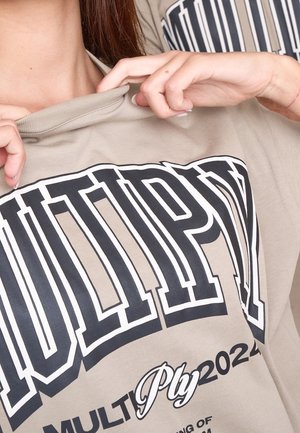 Light beige t-shirt with a rounded neckline, featuring bold black printed text and graphic patterns. Soft fabric texture is visible.