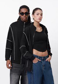Black zip-up jacket with white stitching details, paired with a black crop top, and a model in blue high-waisted jeans.