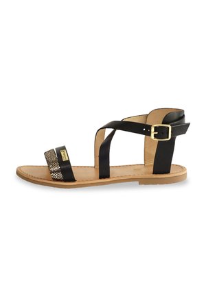 Sandalia de cuero negro con tiras cruzadas, una banda texturizada con acentos dorados y una suela marrón clara. Cuenta con un broche en la correa del tobillo.