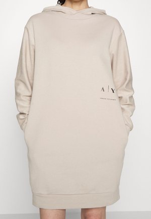 Beige hupparimekko, jossa on pitkät hihat, etukangaroo-taskut ja musta logo vasemmassa alakulmassa. Pehmeä, sileä kangas ja rento istuvuus.