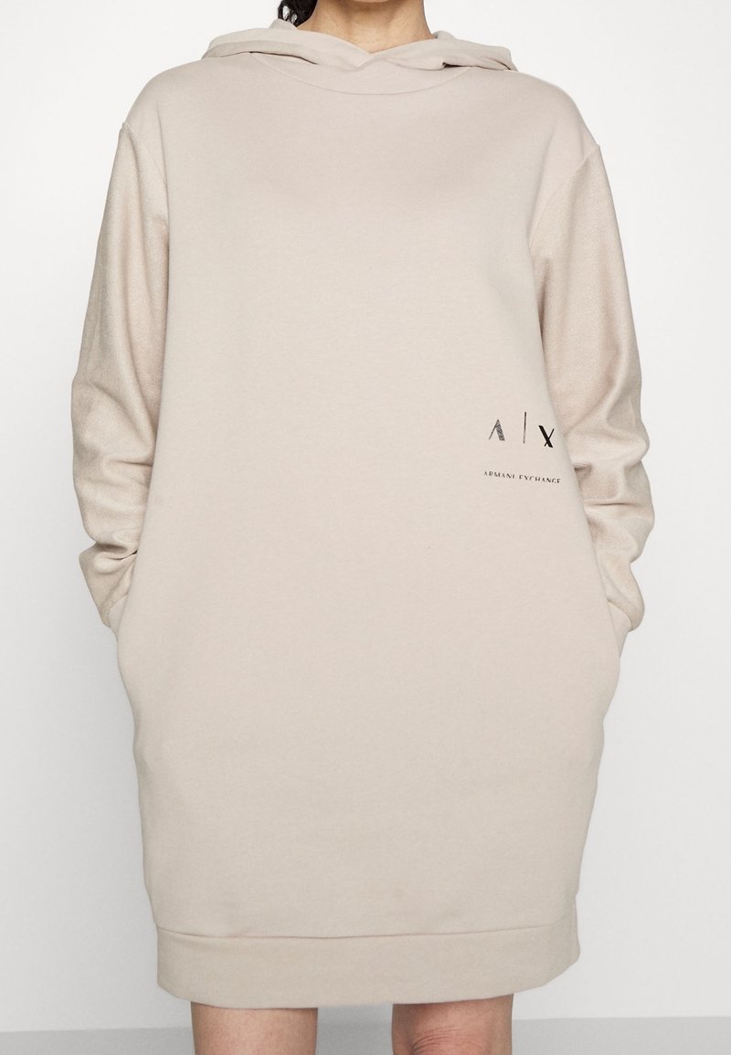 Robe sweat à capuche beige avec manches longues, poches kangourou à l'avant et détail logo noir en bas à gauche. Tissu doux et lisse avec une coupe décontractée.