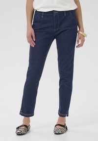 Jean en denim bleu foncé à coupe slim, avec des coutures jaunes contrastantes et un design à cinq poches, associés à des ballerines rayées noir et blanc.