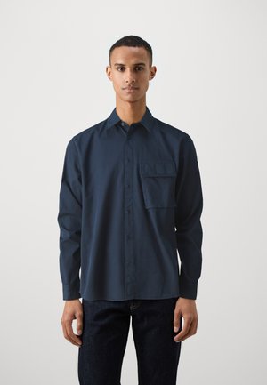Mørkeblå langermet skjorte med knapper laget av bomull, med en lomme på venstre bryst og klassisk krage. Brukt med mørke jeans.