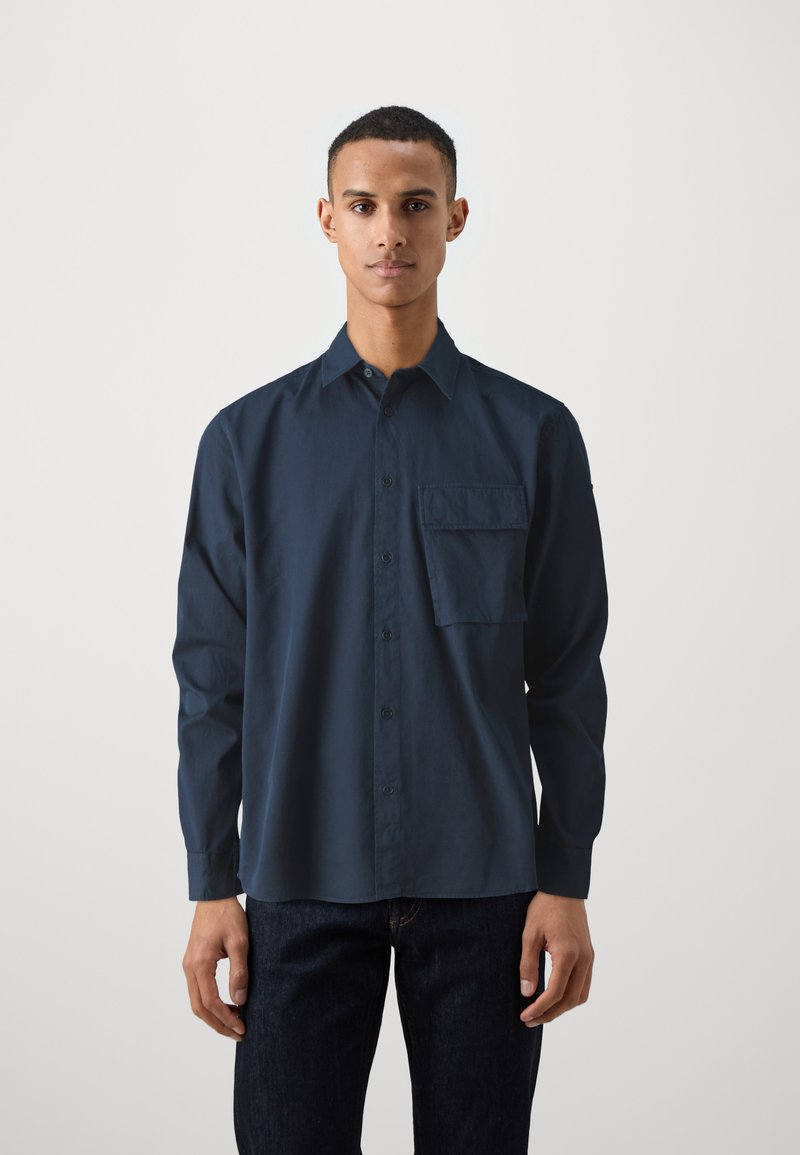 Mørkeblå langermet skjorte med knapper laget av bomull, med en lomme på venstre bryst og klassisk krage. Brukt med mørke jeans.