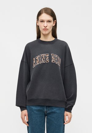 Γυναίκα με ίσια καστανά μαλλιά φοράει φαρδιά σκουρόχρωμη μπλούζα με το κείμενο «ANINE BING» και μπλε τζιν, στέκεται μπροστά σε απλό φωτεινό φόντο.