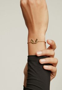 Pulsera de oro con un diseño de chevron texturizado, con dos extremos puntiagudos. Lleva puesta en una muñeca, con una cadena fina y un acabado suave.